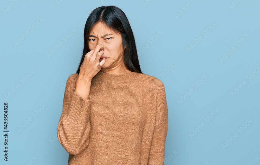Beautiful young asian woman wearing casual winter sweater smelling something stinky and disgusting, intolerable smell, holding breath with fingers on nose. bad smell Beautiful young asian woman wearing casual winter sweater smelling something stinky and disgusting, intolerable smell, holding breath with fingers on nose. bad smell