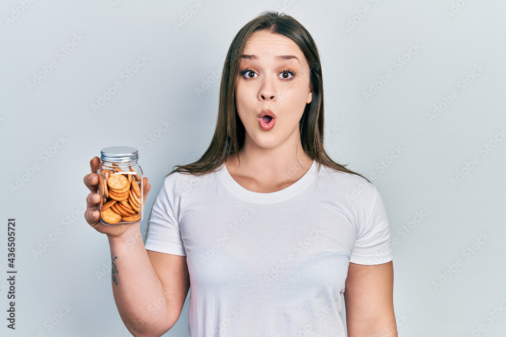 Young hispanic girl holding salty biscuits jar scared and amazed with open mouth for surprise, disbelief face Young hispanic girl holding salty biscuits jar scared and amazed with open mouth for surprise, disbelief face