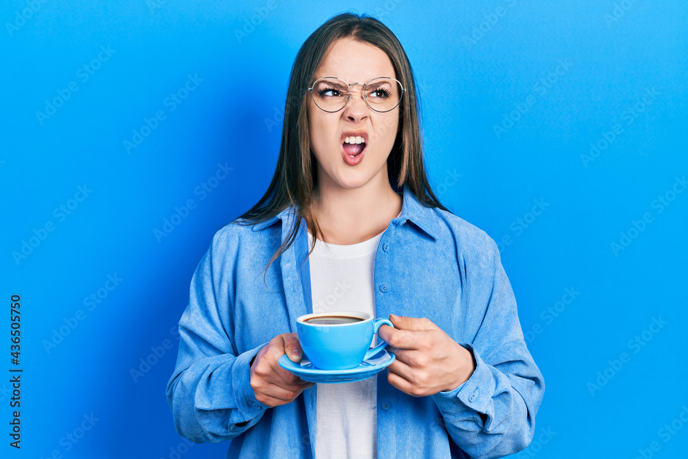 Young hispanic girl drinking a cup of coffee angry and mad screaming frustrated and furious, shouting with anger. rage and aggressive concept. Young hispanic girl drinking a cup of coffee angry and mad screaming frustrated and furious, shouting with anger. rage and aggressive concept.