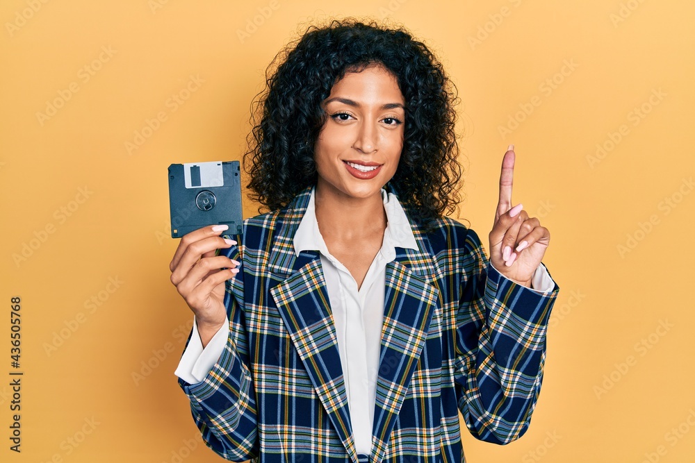 Young latin girl holding floppy disk surprised with an idea or question pointing finger with happy face, number one Young latin girl holding floppy disk surprised with an idea or question pointing finger with happy face, number one