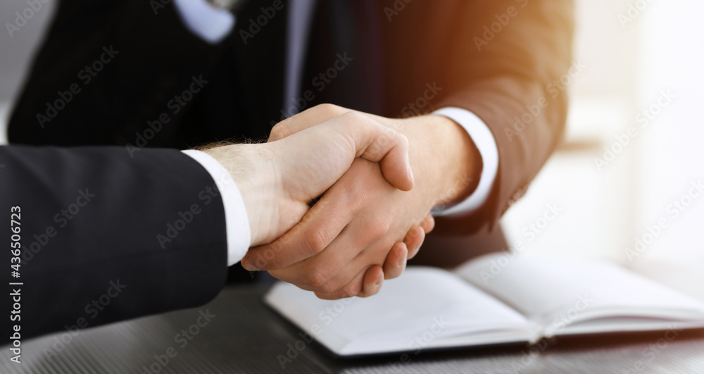 Fototapeta premium Businessman shaking hands with his colleague above the glass desk in sunny modern office, close-up. Unknown business people at meeting