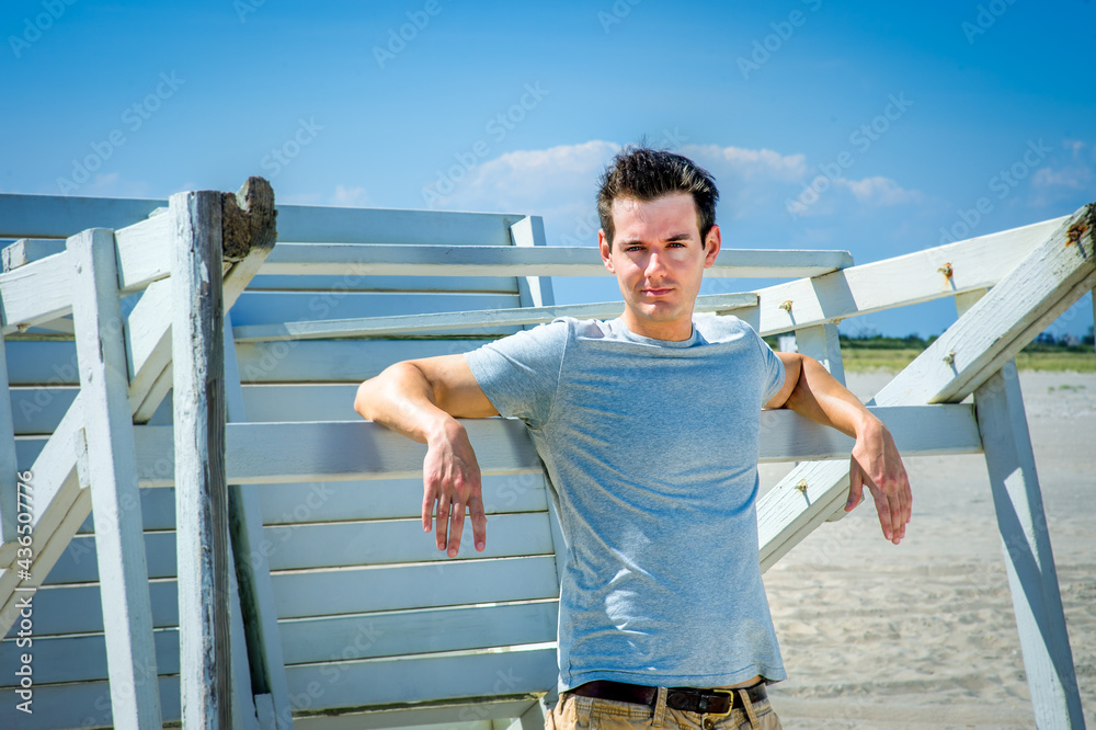Man waiting for you. Wearing a gray t-shirt and arms resting on a wooden stick, a handsome young man is standing by a wooden structure on the beach, narrowing eyes, charmingly looking at you..