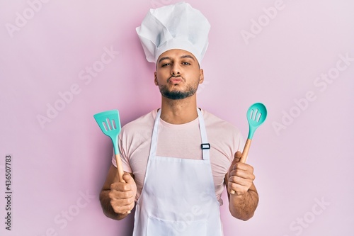 Young arab man wearing professional cook apron and hat holding spoon looking at the camera blowing a kiss being lovely and sexy. love expression. photo