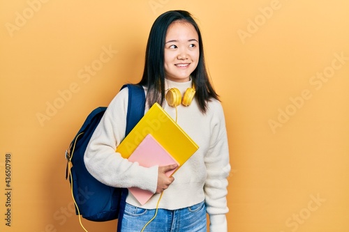 Young chinese girl holding student backpack and books smiling looking to the side and staring away thinking.