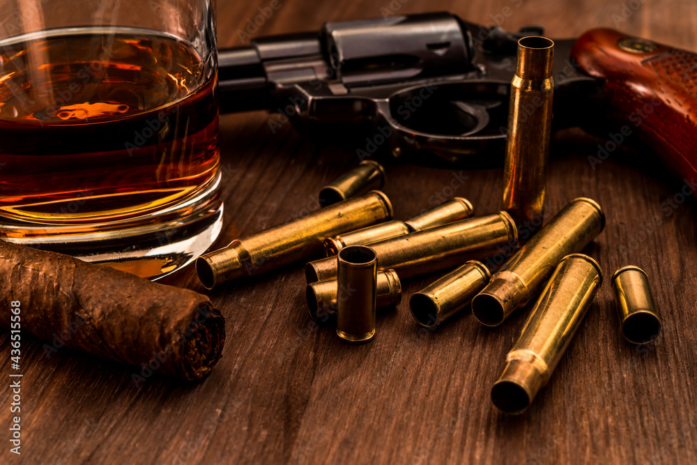 Empty shells from the weapons with glass of whiskey and revolver with cuban cigar on a wooden table