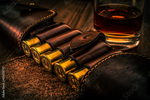 Hunting ammunition 12 gauge in leather bandolier with glass of whiskey on a wooden table. Focus on the cartridges