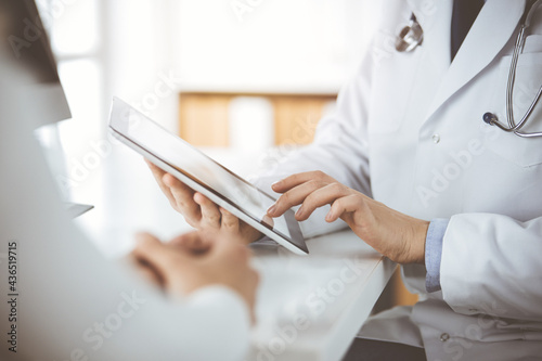 Unknown male doctor and patient woman discussing current health examination while sitting in sunny clinic and using tablet computer. Perfect medical service in hospital