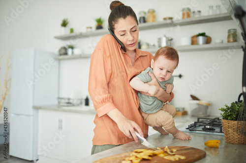 Fotografija Waist up portrait of busy mother holding baby looking at camera while cooking in