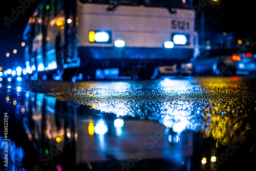 Rainy night in the big city, city bus comes to a halt. View from puddles on the pavement level