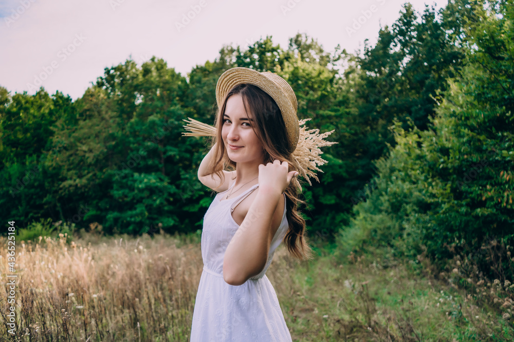 Young woman in a straw hat on a background of tall herbs. Beautiful girl in a dress with a bouquet of dried flowers in the field.
