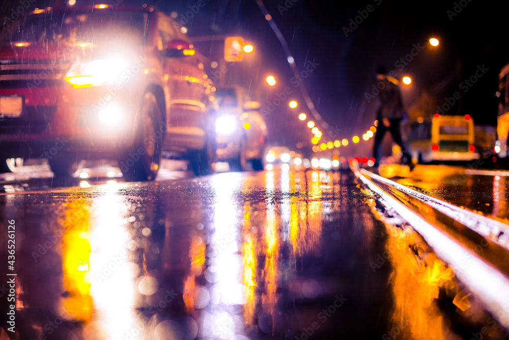 Rainy night in the big city, pedestrians cross the busy intersection. View from the level of a manhole on the pavement Rainy night in the big city, pedestrians cross the busy intersection. View from the level of a manhole on the pavement