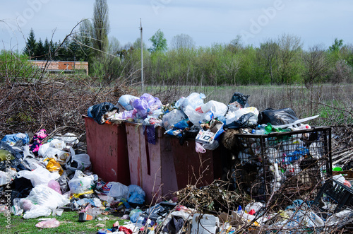 Contamination of nature with human waste. Garbage containers with trash. Overflowing trash cans with pile of rubbish are located outside the village.
