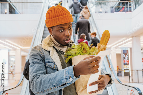 African man with a paper bag of groceries looks surprised and upset at a receipt from a supermarket with high prices against the background of an escalator with customers in the shopping center. The photo