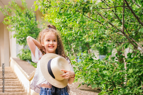 A little girl stands in the park and holds a hat in her hands. The child is happy and laughs.