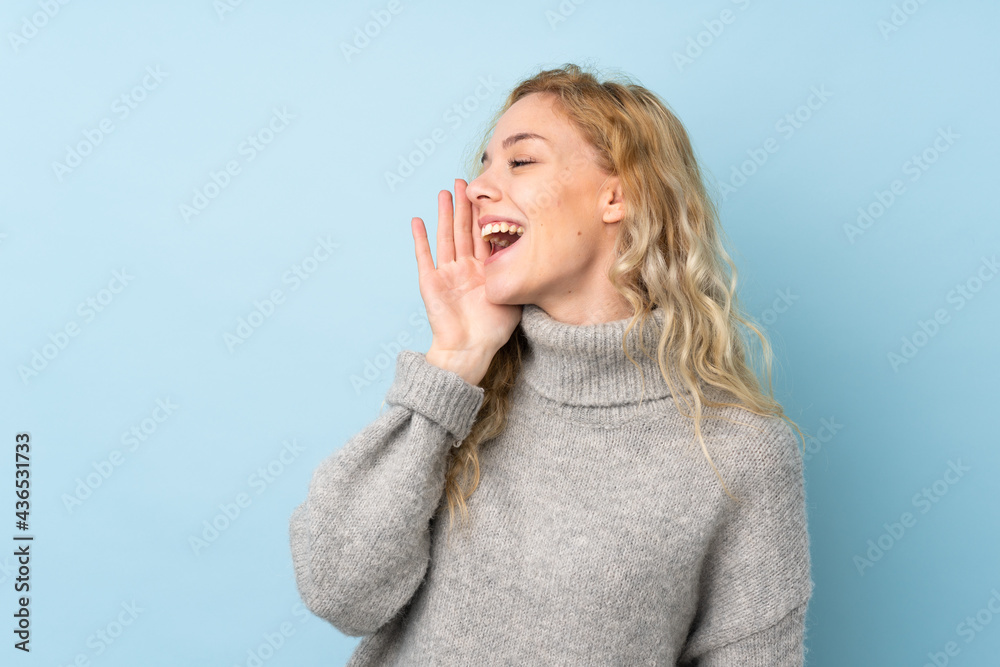 Young blonde woman wearing a sweater isolated on blue background shouting with mouth wide open to the lateral Young blonde woman wearing a sweater isolated on blue background shouting with mouth wide open to the lateral