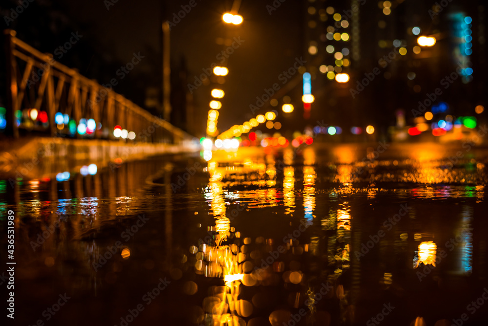Rainy night in the big city, approaching headlights of cars traveling along the avenue.  View from puddles on the pavement level