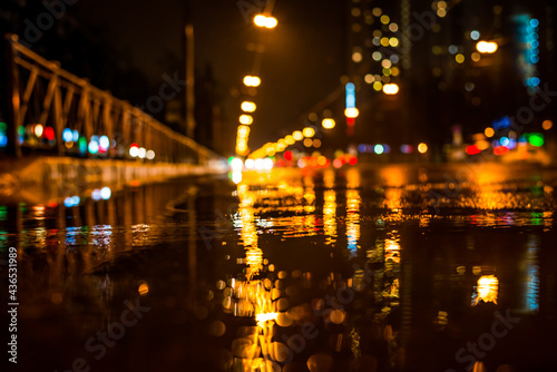 Rainy night in the big city, approaching headlights of cars traveling along the avenue. View from puddles on the pavement level
