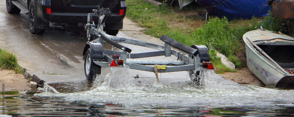 Motor boat launch, SUV car with wet empty boat trailer move on concrete shipway from water at summer day on old boat on shore background
