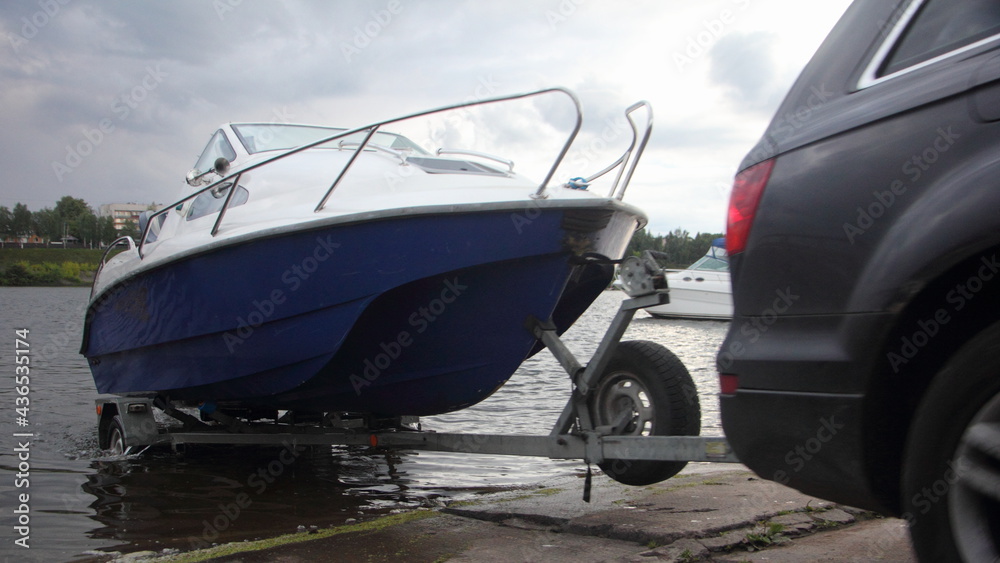 A 4x4 black car pulls blue-white trimaran cabin motor boat on trailer on concrete slipway into the river water, boat launch before active recreation on the river