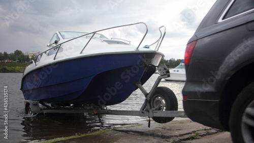 Obraz na płótnie A 4x4 black car pulls blue-white trimaran cabin motor boat on trailer on concrete slipway into the river water, boat launch before active recreation on the river w salonie