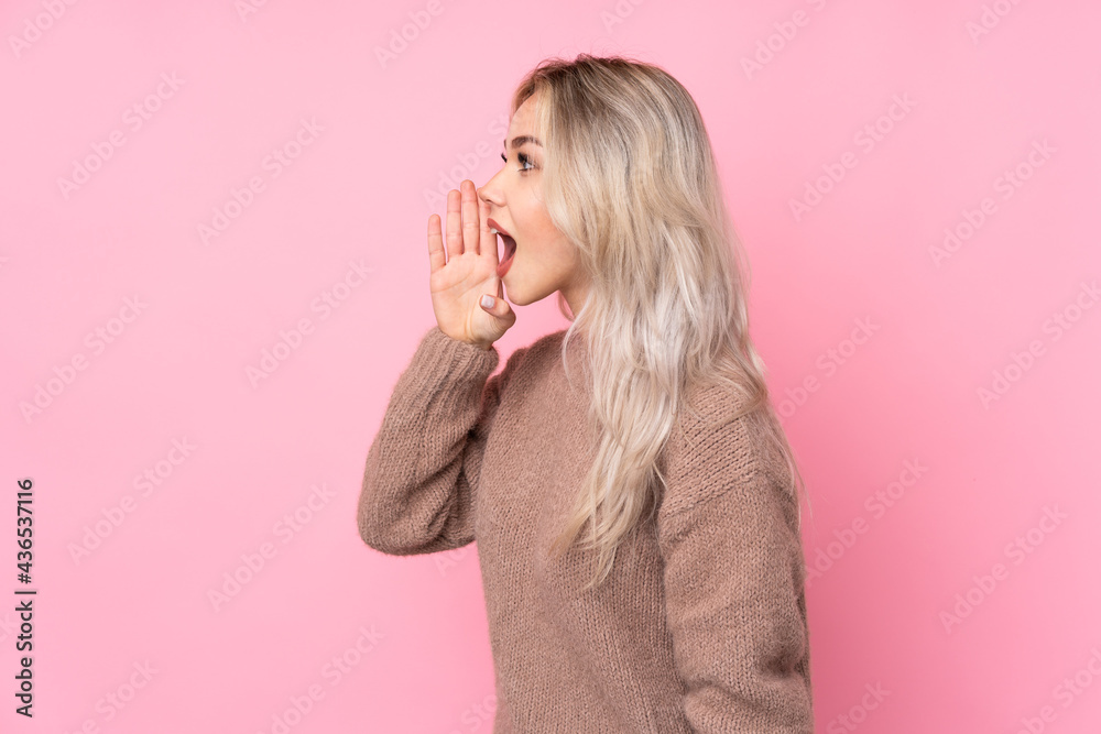Teenager blonde girl wearing a sweater over isolated pink background shouting with mouth wide open to the lateral Teenager blonde girl wearing a sweater over isolated pink background shouting with mouth wide open to the lateral