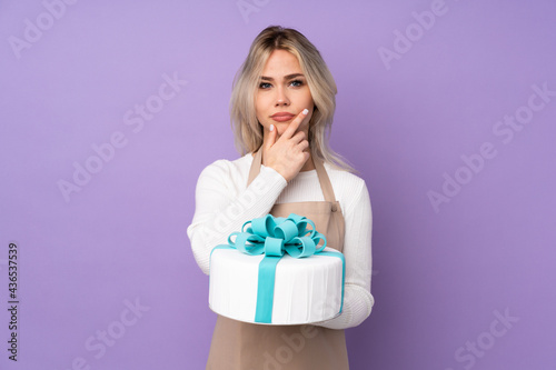 Young pastry chef holding a big cake over isolated purple background thinking an idea