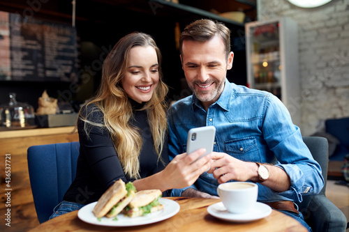 Happy mid adult couple sharing phone at cafe having lunch break
