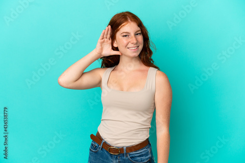 Teenager reddish woman isolated on blue background listening to something by putting hand on the ear