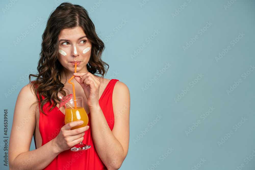 Charming girl drinking orange juice looks away. Girl wears a red, one-piece swimsuit on a blue background Charming girl drinking orange juice looks away. Girl wears a red, one-piece swimsuit on a blue background