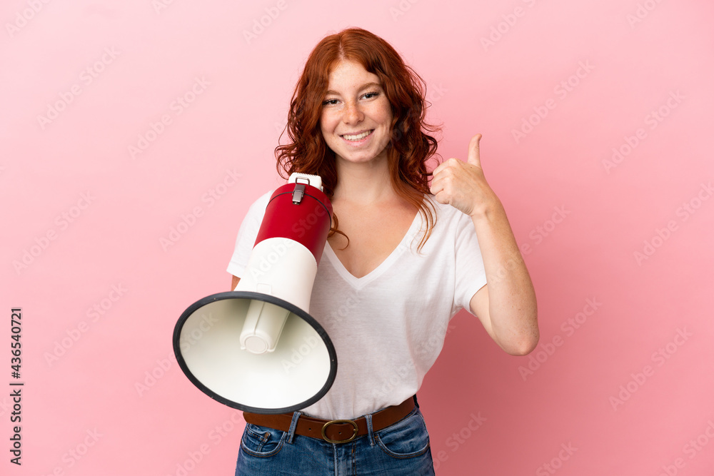 Teenager reddish woman isolated on pink background holding a megaphone with thumb up Teenager reddish woman isolated on pink background holding a megaphone with thumb up