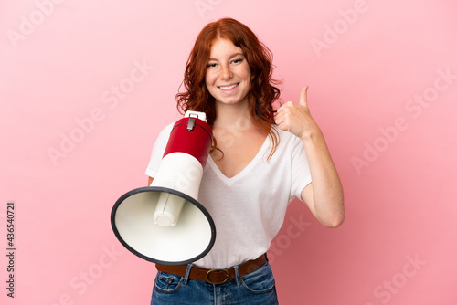 Teenager reddish woman isolated on pink background holding a megaphone with thumb up