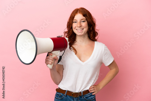Teenager reddish woman isolated on pink background holding a megaphone and smiling