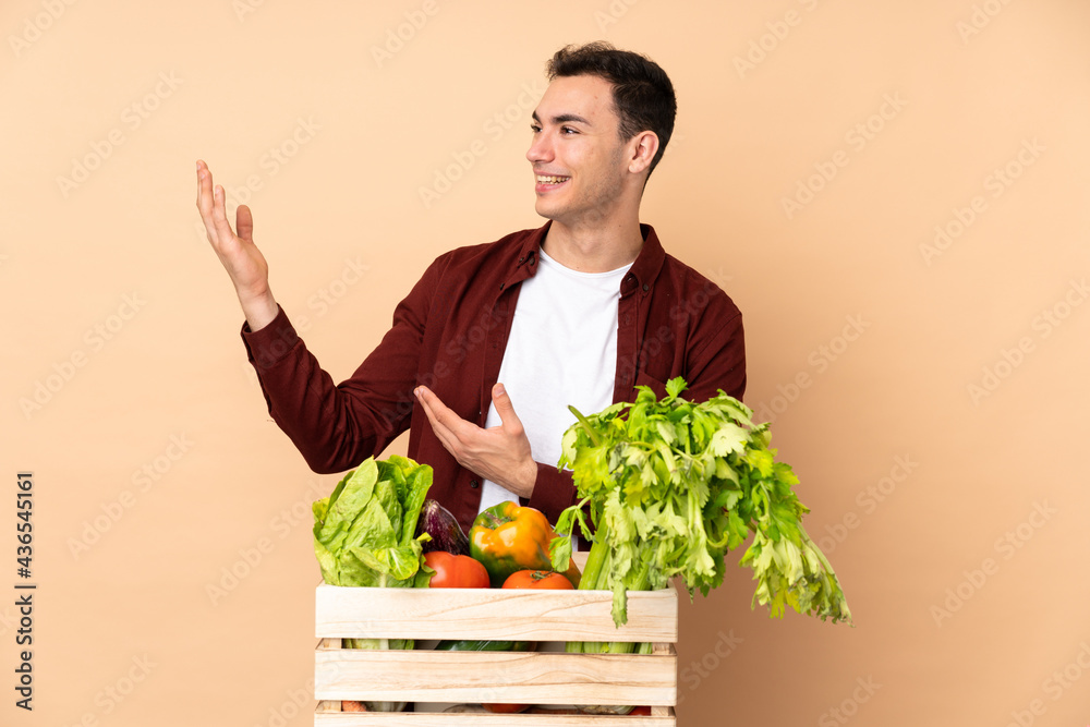 Farmer with freshly picked vegetables in a box isolated on beige background extending hands to the side for inviting to come Farmer with freshly picked vegetables in a box isolated on beige background extending hands to the side for inviting to come