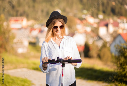 Young blond woman in black hat and sunglasses sets the ride mode on the touch panel. Stylish girl riding on electric scooter in city center. Summer leisure activity. Spending free time outdoors in