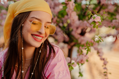 Fashionable happy smiling woman wearing stylish yellow bandana, trendy sunglasses with chain, posing near blooming tree. Spring, summer fashion, lifestyle conception. Copy, empty space for text 