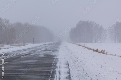 Headlights light of car traveling during the snow hurricane on the highway next to the village in the evening.