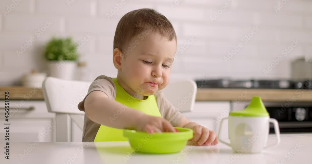 Cute little baby boy sits at a table in the kitchen and eats strawberries from green plate with his hands. Slow motion. Cute little baby boy sits at a table in the kitchen and eats strawberries from green plate with his hands. Slow motion.