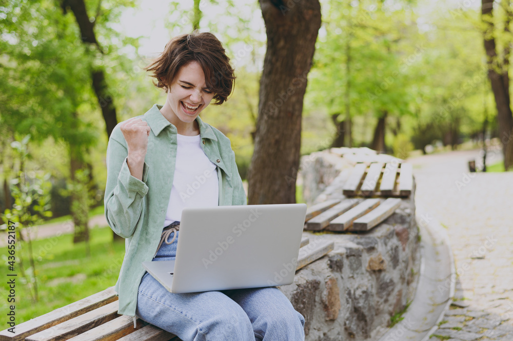 Young student freelancer woman in green jacket jeans sit on bench in spring park outdoors rest use laptop pc computer with wi-fi internet do winner gesture clench fist People urban lifestyle concept