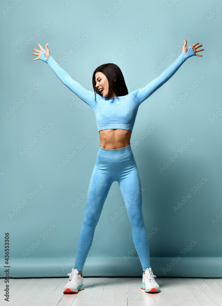Portrait of fitness girl. Woman in fashion sportswear, standing on a blue  background, after a workout. Fit girls with a strong muscular body, raised  her arms up, rejoices in her body Stock