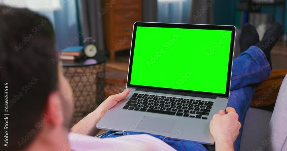 Young Man at Home Uses Green Mock-up Screen Laptop. He is lying On a Couch in His Cozy Living Room. Male student is talking on video chat with friends, doctor or financial agent Young Man at Home Uses Green Mock-up Screen Laptop. He is lying On a Couch in His Cozy Living Room. Male student is talking on video chat with friends, doctor or financial agent