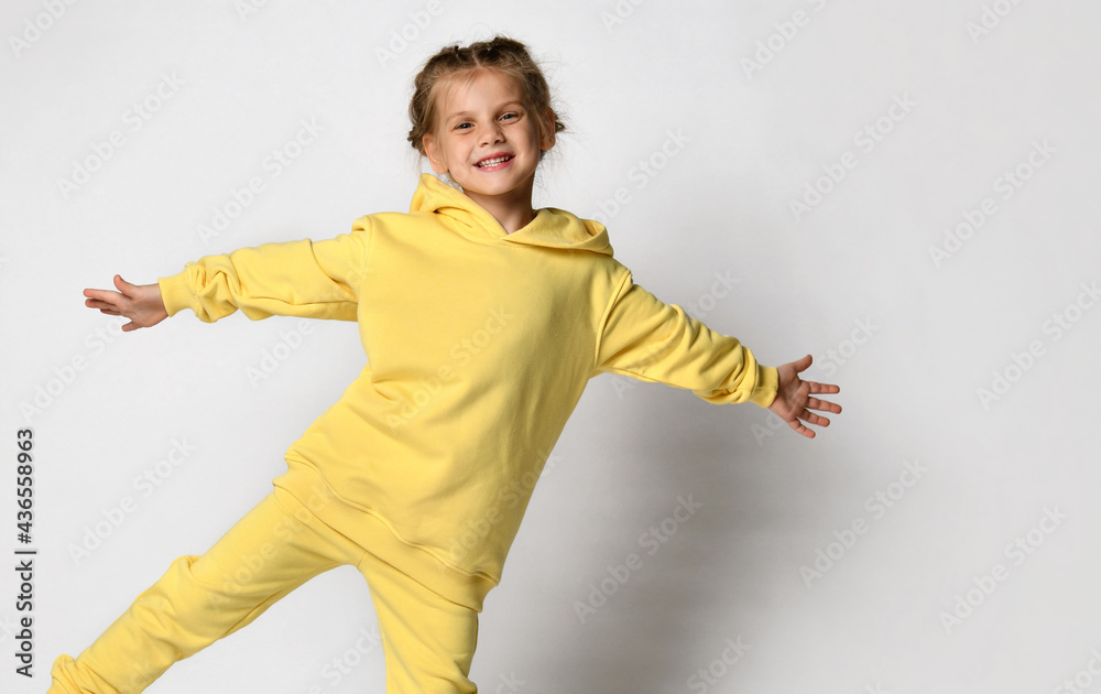 Beautiful little girl having fun with outstretched arms showing off her new tracksuit. Cute child posing in a stylish yellow sports suit on a white background. Concept of stylish sportswear.
