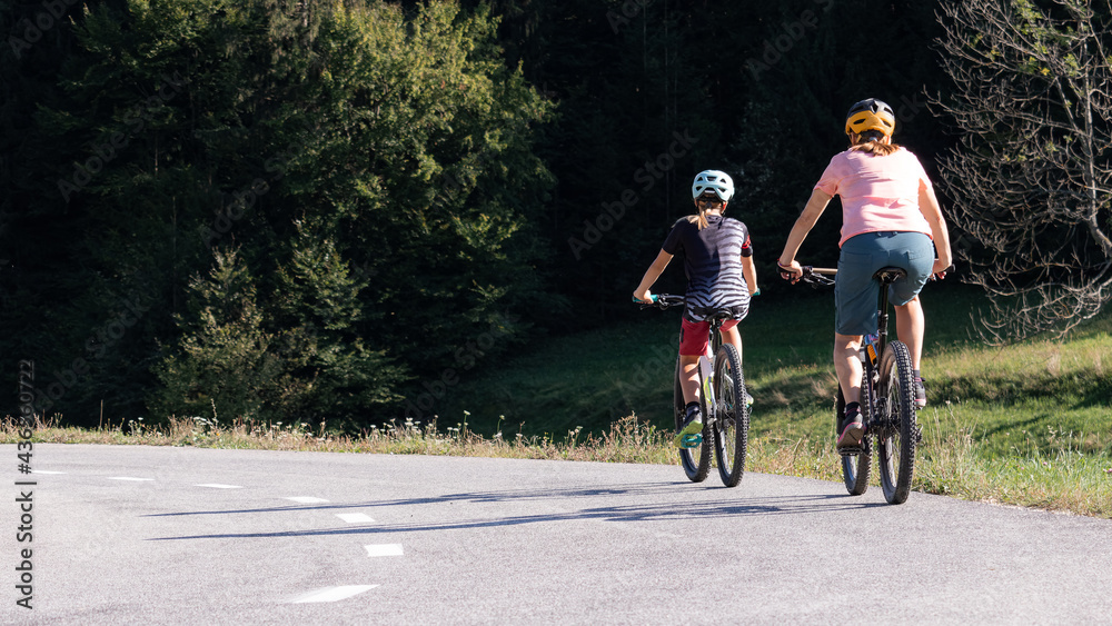 Woman and girl child riding mountain bikes on a country road in nature on a bright sunny summer day.