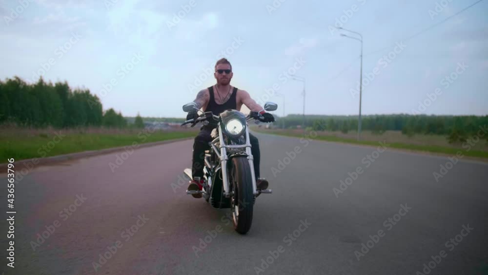Stylish biker with tattoos rides a motorcycle on a country road at sunset Stylish biker with tattoos rides a motorcycle on a country road at sunset