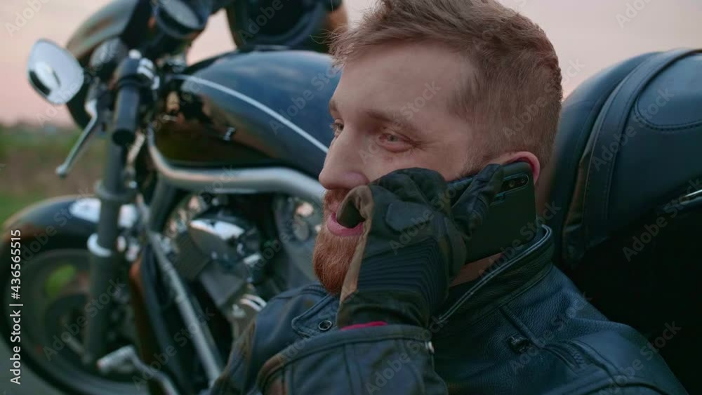 A middle-aged male biker sits by a motorcycle and talks on his mobile phone A middle-aged male biker sits by a motorcycle and talks on his mobile phone