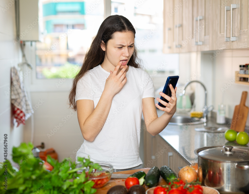 Portrait of attractive pretty girl reading recipe online on the phone in the kitchen