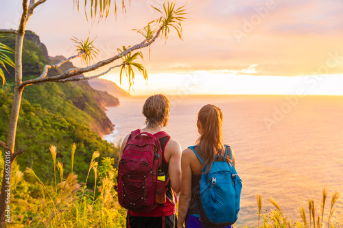 Hawaii hiking hikers hiking on Kalalau trail watching sunset from Na Pali Coast. Tourists couple with backpacks walking outdoor in Kauai island. Summer travel adventure active lifestyle. photo