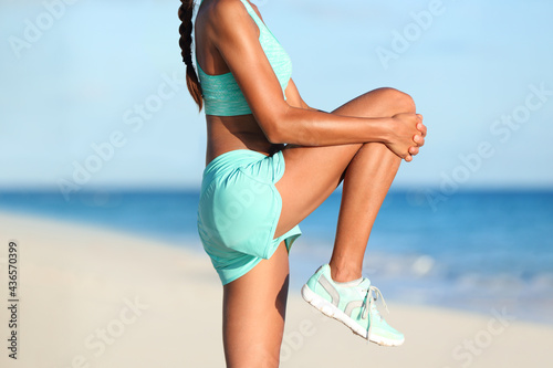 Fitness stretches woman runner stretching glute muscle with dynamic high knee pull stretch. Athlete getting ready to run doing leg muscles warm-up standing exercise on beach pre-workout. Body closeup. photo