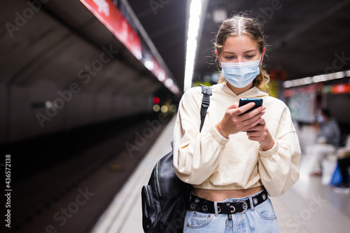 Portrait of a confident girl in a protective mask walking on a subway station platform during a pandemic and texting ..with friends on a mobile phone while waiting for a train photo