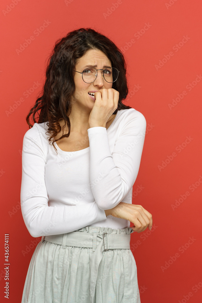 Worried female in eyeglasses look scared and nervous bite nails in anxiety. Troubled frightened young woman look in camera over red studio background. Stressed girl anxious think of problem or mistake Worried female in eyeglasses look scared and nervous bite nails in anxiety. Troubled frightened young woman look in camera over red studio background. Stressed girl anxious think of problem or mistake