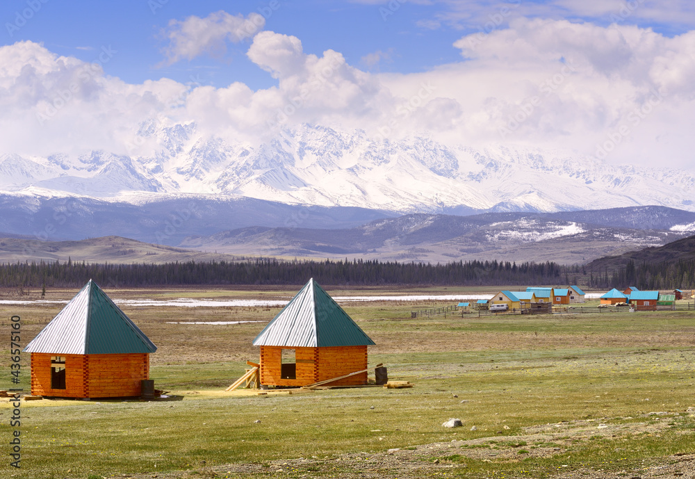 Kurai steppe in spring. Tourist houses with a conical roof on the background of the snow-capped peaks of the Northern Chui ridge under a cloudy blue sky. Gorny Altai, Siberia, Russia